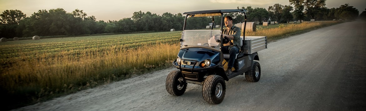 Man driving a blue lifted Cushman Hauler Elite down a gravel road, surrounded by a field spotted with bales …
